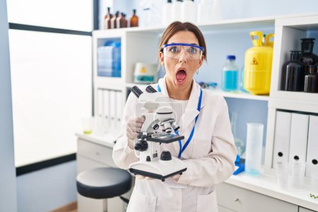 Photo for Young brunette woman working at scientist laboratory with microscope afraid and shocked with surprise and amazed expression, fear and excited face. - Royalty Free Image