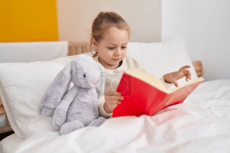 Photo for Adorable blonde girl reading book sitting on bed at bedroom - Royalty Free Image