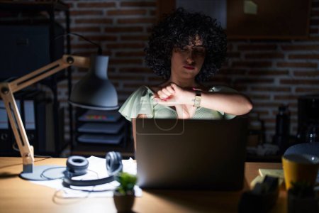 Téléchargez les photos : Young brunette woman with curly hair working at the office at night checking the time on wrist watch, relaxed and confident - en image libre de droit