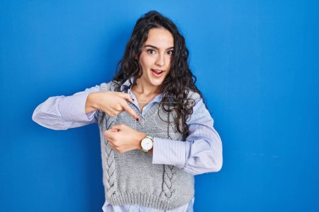 Foto de Young brunette woman standing over blue background in hurry pointing to watch time, impatience, upset and angry for deadline delay - Imagen libre de derechos