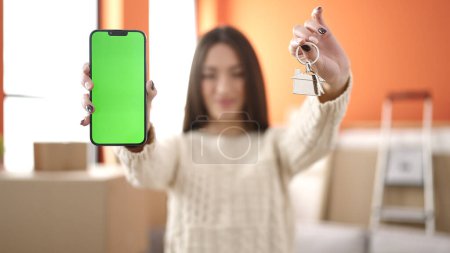 Photo for Young beautiful hispanic woman smiling confident holding key showing screen smartpho at new home - Royalty Free Image