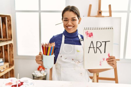 Photo for Young latin woman holding art notebook and cup with pencils at art studio - Royalty Free Image