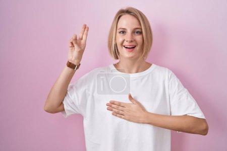 Téléchargez les photos : Jeune femme caucasienne debout sur fond rose souriant jurant avec la main sur la poitrine et les doigts vers le haut, faisant un serment de fidélité promesse - en image libre de droit
