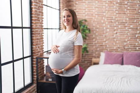 Foto de Joven embarazada sonriendo confiado tocando vientre de pie en el dormitorio - Imagen libre de derechos