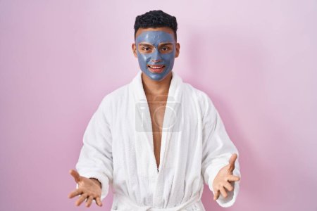 Foto de Young hispanic man wearing beauty face mask and bath robe smiling cheerful with open arms as friendly welcome, positive and confident greetings - Imagen libre de derechos