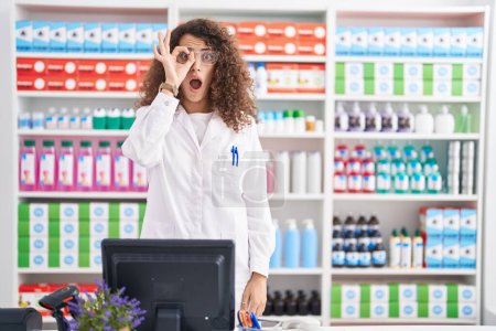Téléchargez les photos : Hispanic woman with curly hair working at pharmacy drugstore doing ok gesture shocked with surprised face, eye looking through fingers. unbelieving expression. - en image libre de droit