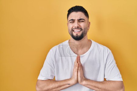 Photo for Young handsome man wearing casual t shirt over yellow background begging and praying with hands together with hope expression on face very emotional and worried. begging. - Royalty Free Image