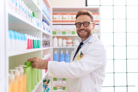 Photo for Middle age man pharmacist smiling confident holding product on shelving at pharmacy - Royalty Free Image