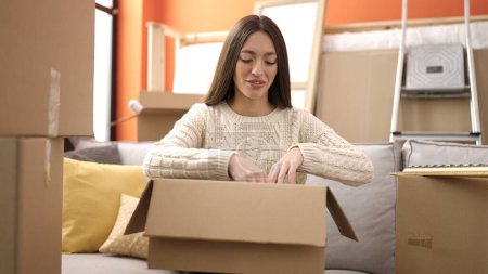 Photo for Young beautiful hispanic woman smiling confident unpacking cardboard box at new home - Royalty Free Image