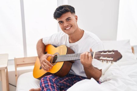 Photo for Young hispanic man playing classical guitar sitting on bed at bedroom - Royalty Free Image