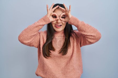 Téléchargez les photos : Young latin woman standing over blue background doing ok gesture like binoculars sticking tongue out, eyes looking through fingers. crazy expression. - en image libre de droit