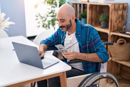Téléchargez les photos : Jeune homme hispanique utilisant ordinateur portable holdig dollars assis sur fauteuil roulant à la maison - en image libre de droit