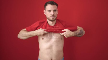 Photo for Young caucasian man standing with serious expression taking t shirt off over isolated red background - Royalty Free Image