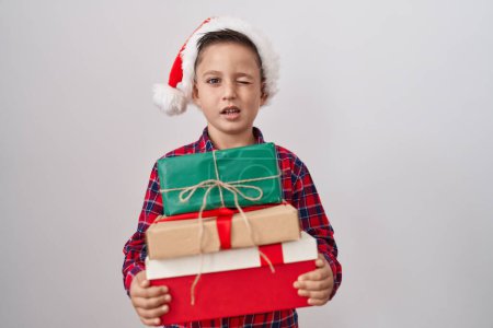 Foto de Pequeño niño hispano con sombrero de navidad sosteniendo regalos guiñando el ojo mirando a la cámara con expresión sexy, cara alegre y feliz. - Imagen libre de derechos