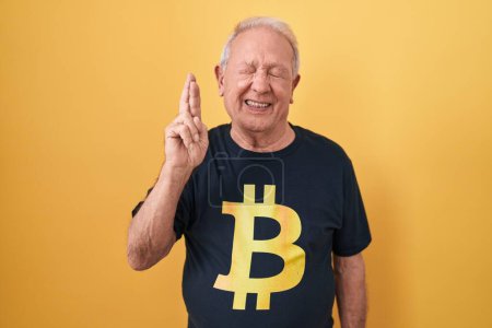 Téléchargez les photos : Senior man with grey hair wearing bitcoin t shirt gesturing finger crossed smiling with hope and eyes closed. luck and superstitious concept. - en image libre de droit