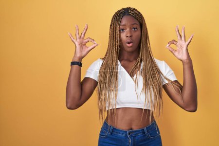 Photo for African american woman with braided hair standing over yellow background looking surprised and shocked doing ok approval symbol with fingers. crazy expression - Royalty Free Image