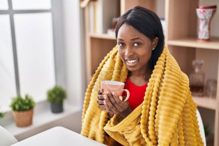 Téléchargez les photos : Jeune femme afro-américaine buvant du café assis sur la table à la maison - en image libre de droit