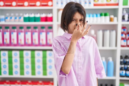 Foto de Young girl at pharmacy drugstore smelling something stinky and disgusting, intolerable smell, holding breath with fingers on nose. bad smell - Imagen libre de derechos