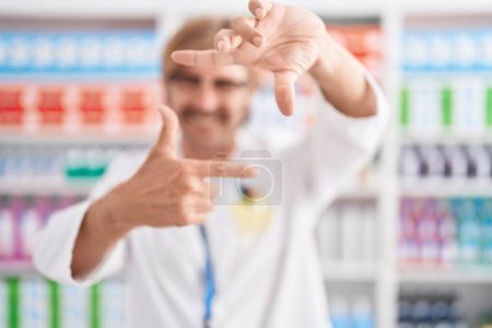 Téléchargez les photos : Homme caucasien avec moustache travaillant à la pharmacie pharmacie souriant faisant cadre avec les mains et les doigts avec le visage heureux. concept de créativité et photographie. - en image libre de droit