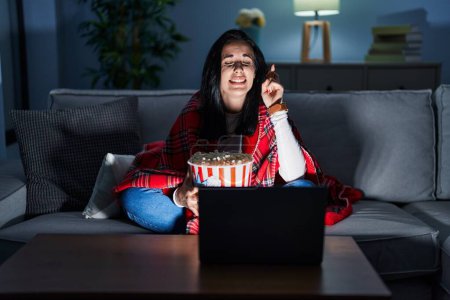 Foto de Hispanic woman eating popcorn watching a movie on the sofa gesturing finger crossed smiling with hope and eyes closed. luck and superstitious concept. - Imagen libre de derechos