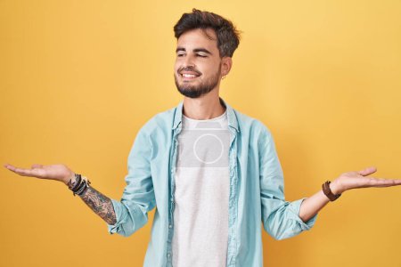 Téléchargez les photos : Young hispanic man with tattoos standing over yellow background smiling showing both hands open palms, presenting and advertising comparison and balance - en image libre de droit