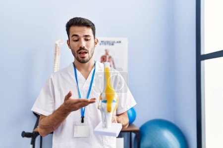 Téléchargez les photos : Jeune homme hispanique portant l'uniforme de physiothérapeute tenant le modèle anatomique du genou à la clinique de réadaptation - en image libre de droit