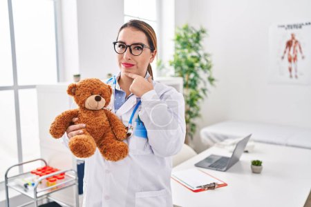 Photo for Young hispanic pediatrician woman holding teddy bear at the clinic serious face thinking about question with hand on chin, thoughtful about confusing idea - Royalty Free Image