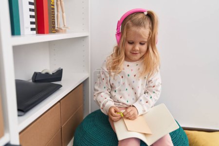 Foto de Adorable chica rubia estudiante escuchando música lectura libro en biblioteca escuela - Imagen libre de derechos