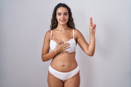 Foto de Mujer hispana joven usando lencería blanca sonriendo jurando con la mano en el pecho y los dedos hacia arriba, haciendo un juramento de lealtad promesa - Imagen libre de derechos