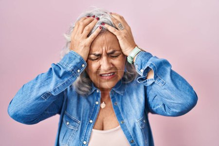 Foto de Mujer de mediana edad con el pelo gris de pie sobre el fondo rosa que sufre de dolor de cabeza desesperada y estresada porque el dolor y la migraña. manos en la cabeza. - Imagen libre de derechos