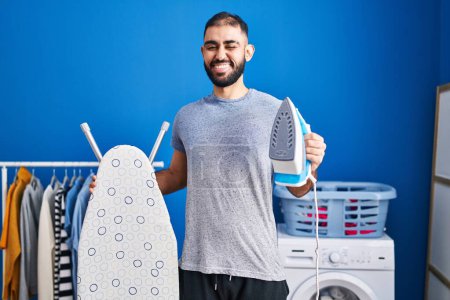 Foto de Middle east man with beard holding electric iron winking looking at the camera with sexy expression, cheerful and happy face. - Imagen libre de derechos