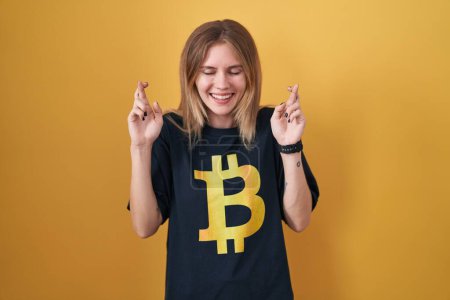Foto de Mujer rubia caucásica vistiendo camiseta bitcoin gesto dedo cruzado sonriendo con esperanza y los ojos cerrados. suerte y concepto supersticioso. - Imagen libre de derechos