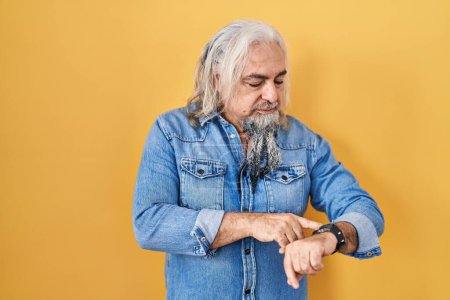 Téléchargez les photos : Homme d'âge moyen avec les cheveux gris debout sur fond jaune vérifier l'heure sur montre-bracelet, détendu et confiant - en image libre de droit
