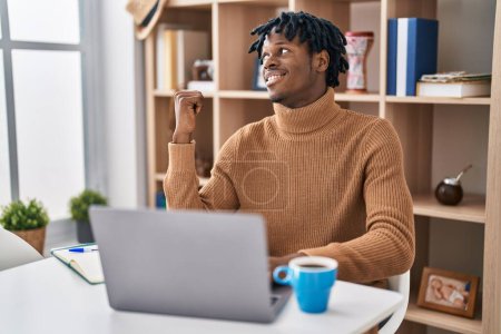 Téléchargez les photos : Jeune homme africain avec dreadlocks travailler en utilisant ordinateur portable souriant avec le visage heureux regardant et pointant vers le côté avec le pouce vers le haut. - en image libre de droit
