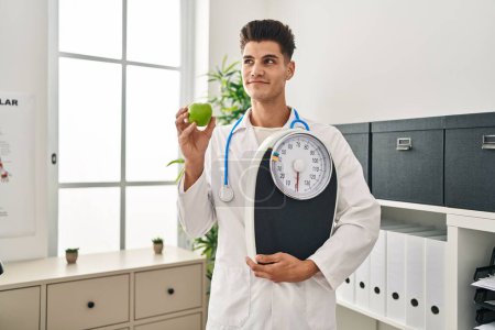 Téléchargez les photos : Young hispanic doctor man holding scale at dietitian clinic smiling looking to the side and staring away thinking. - en image libre de droit