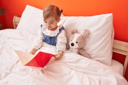 Foto de Adorable rubio niño leyendo libro sentado en la cama con oso de peluche en el dormitorio - Imagen libre de derechos