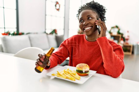 Photo for African american woman eating hamburger talking on smartphone sitting on table by christmas tree at home - Royalty Free Image