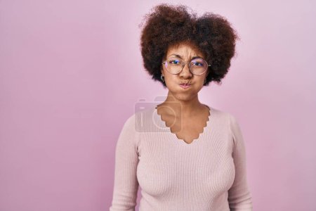 Foto de Joven mujer afroamericana de pie sobre fondo rosa hinchando mejillas con cara divertida. boca hinchada de aire, expresión loca. - Imagen libre de derechos