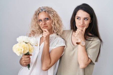 Photo for Mother and daughter holding bouquet of white flowers looking confident at the camera smiling with crossed arms and hand raised on chin. thinking positive. - Royalty Free Image