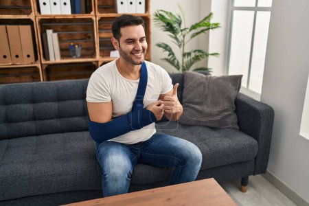 Foto de Young hispanic man with beard wearing arm on sling sitting at therapy consult smiling happy and positive, thumb up doing excellent and approval sign - Imagen libre de derechos