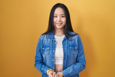 Téléchargez les photos : Jeune femme asiatique debout sur fond jaune avec les mains jointes et les doigts croisés souriant détendu et joyeux. succès et optimisme - en image libre de droit