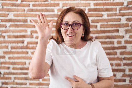 Foto de Mujer mayor con gafas de pie sobre la pared de ladrillos renunciando a decir hola feliz y sonriente, gesto de bienvenida amistoso - Imagen libre de derechos