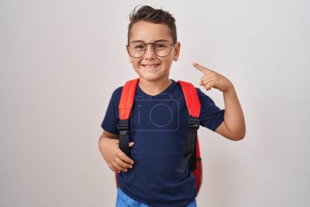 Téléchargez les photos : Petit garçon hispanique portant des lunettes et sac à dos étudiant souriant heureux pointant avec la main et le doigt - en image libre de droit
