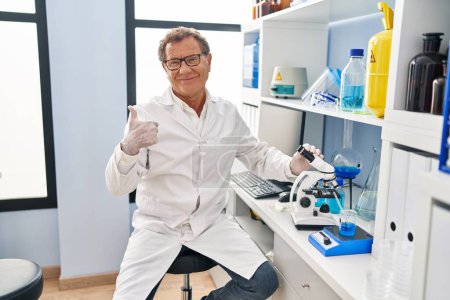 Photo for Senior man working at scientist laboratory smiling happy and positive, thumb up doing excellent and approval sign - Royalty Free Image