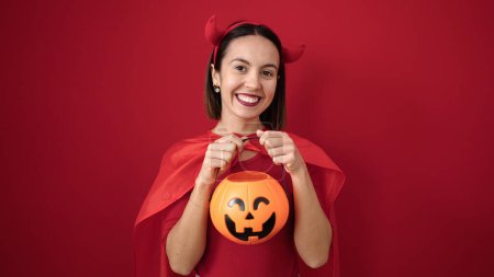 Téléchargez les photos : Young beautiful hispanic woman wearing devil costume holding halloween pumpkin basket over isolated red background - en image libre de droit