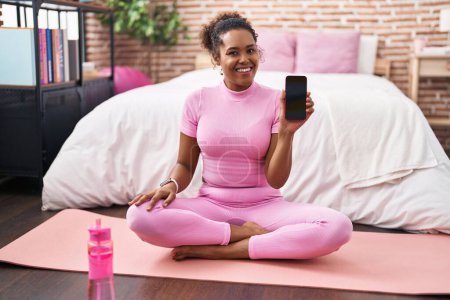 Téléchargez les photos : Jeune femme hispanique avec les cheveux bouclés assis sur le tapis de yoga dans la chambre à coucher regardant positif et heureux debout et souriant avec un sourire confiant montrant les dents - en image libre de droit