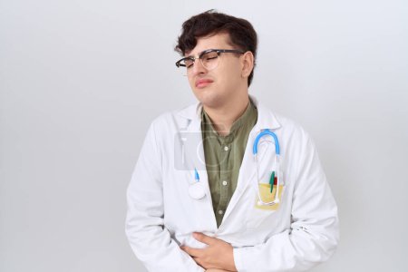 Téléchargez les photos : Young non binary man wearing doctor uniform and stethoscope with hand on stomach because nausea, painful disease feeling unwell. ache concept. - en image libre de droit