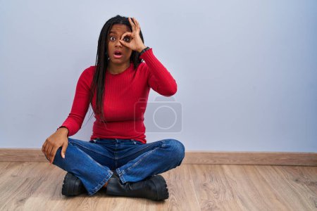 Foto de Joven afroamericano con trenzas sentado en el suelo en casa haciendo buen gesto conmocionado con la cara sorprendida, los ojos mirando a través de los dedos. expresión incrédula. - Imagen libre de derechos