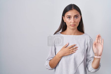 Foto de Young hispanic woman standing over white background swearing with hand on chest and open palm, making a loyalty promise oath - Imagen libre de derechos