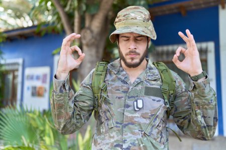 Téléchargez les photos : Jeune homme hispanique portant un uniforme de camouflage de l'armée à l'extérieur se détendre et sourire les yeux fermés en faisant geste de méditation avec les doigts. concept de yoga. - en image libre de droit